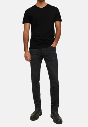 Schwarzes V-Ausschnitt-T-Shirt, dunkle Jeans und schwarze Ankle-Boots. Slim-Fit mit glatter Textur und einem dezenten Glanz auf den Jeans.