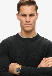 Svart tröja i waffle-knits med rund hals och långa ärmar, som bärs över en grå undertröja. En brun läderklocka med blå urtavla är synlig.