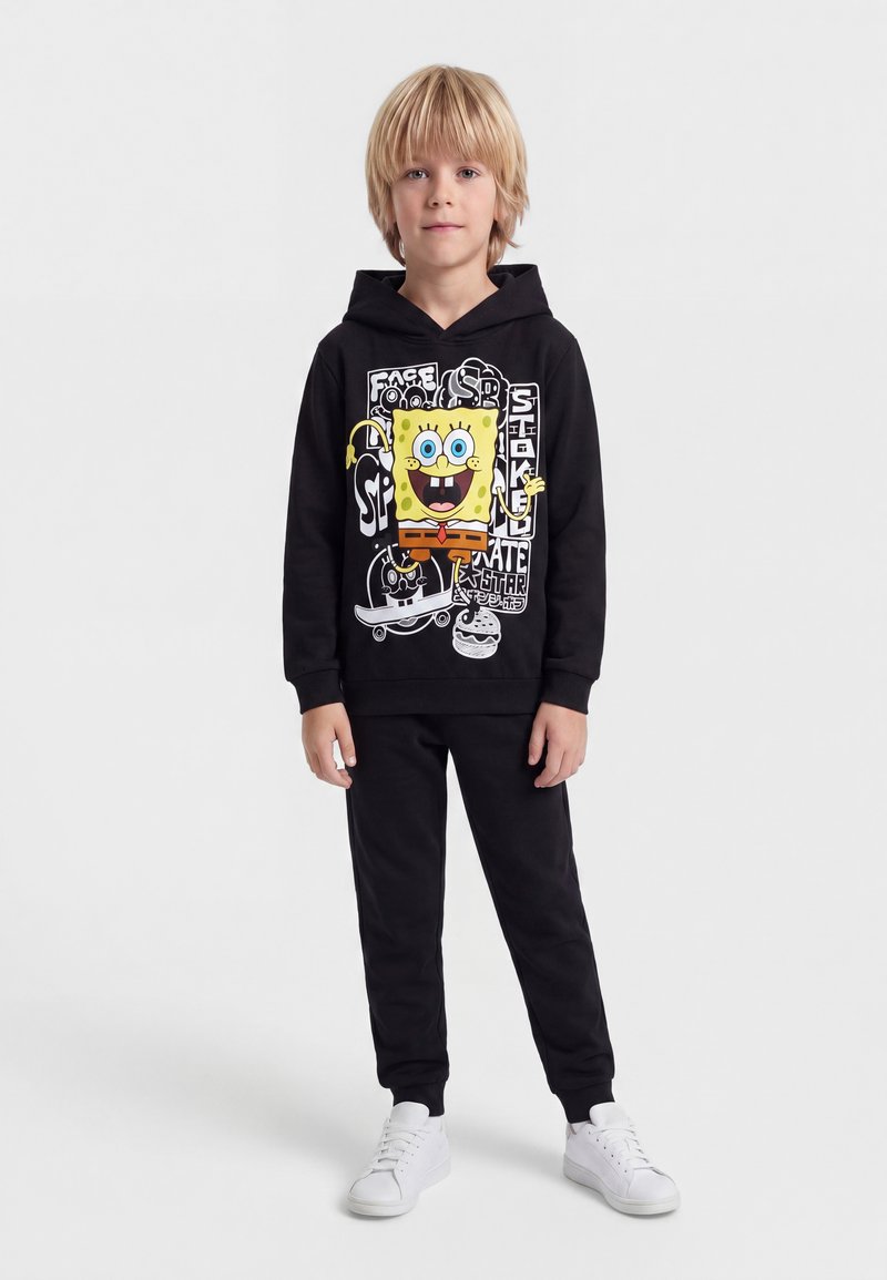 Garçon blond portant un sweat à capuche noir avec un motif coloré de Bob l'éponge, pantalon noir et baskets blanches, debout devant un fond uni.