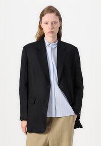 Blazer noir à bouton unique, avec des épaules structurées et des poches avant, porté sur une chemise à rayures bleu clair et un pantalon beige.