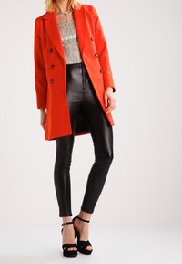 Manteau rouge à double boutonnage sur un top argenté pailleté, leggings noirs en simili cuir et talons aiguilles noirs. Textures lisses et lignes nettes.