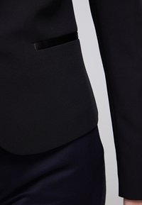 Close-up of a tailored black blazer sleeve and pocket detail worn over dark pants on a gray background.