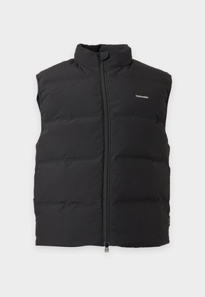 Modèle masculin portant une doudoune noire par-dessus une chemise manches longues bleu marine, un pantalon gris à jambes larges et des baskets marron, debout devant un fond blanc.