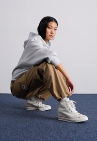 Grå hettegenser, beige joggebukser med elastiske mansjetter, og hvite høye sneakers. Glatt stoff med avslappet passform og synlig merke-patch.