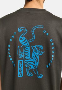Grijze T-shirt met een blauwe afbeelding van een tijger in een rechtopstaande positie, met de tekst "DE BALANS VAN TEGENSTELLINGEN" en "RVCA" eronder.