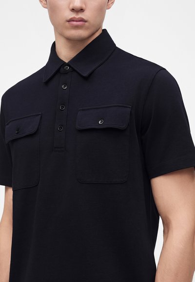 Homme portant une chemise noire à manches courtes avec un col et une patte de boutonnage à quatre boutons, avec deux poches poitrine boutonnées.