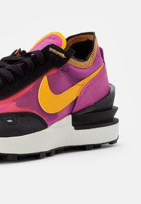 Tenisky s horní částí z fialové síťoviny, černými semišovými akcenty a žlutým logem Nike. Mají robustní bílou a černou podrážku s texturovaným vzorkem.