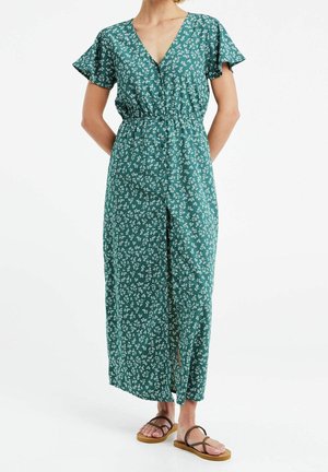 Femme portant une robe maxi verte à motifs floraux avec manches courtes et col en V, associée à des sandales plates marron, debout sur fond blanc.