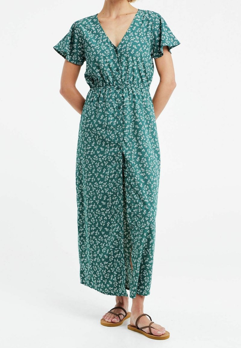 Femme portant une robe maxi verte à motifs floraux avec manches courtes et col en V, associée à des sandales plates marron, debout sur fond blanc.