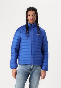 Blau gepolsterte Jacke mit hohem Kragen, horizontalen Nähten und einem Reißverschluss vorne. Sie verfügt über ein Logo auf der Brust. Getragen mit hellen Jeans.