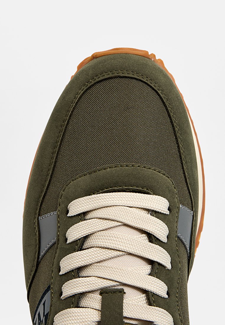 Olivgrüne Sneaker mit strukturiertem Textil-Obermaterial, cremefarbenen Schnürsenkeln, grauem Akzentstreifen und brauner Gummisohle. Verfügt über eine gepolsterte Lasche für zusätzlichen Komfort.