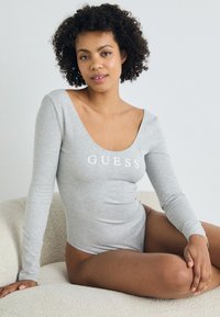 Šedý bodysuit s dlouhým rukávem a kulatým výstřihem, na hrudi s bílým logem "GUESS", vyrobený z hladkého, elastického materiálu a úzkého střihu.