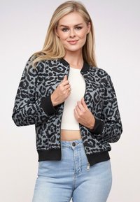 Blondynka w czarno-szarym kurtce z nadrukiem w panterę na zamek, założonej na białą bluzkę typu crop top i jasnoniebieskie dżinsy.
