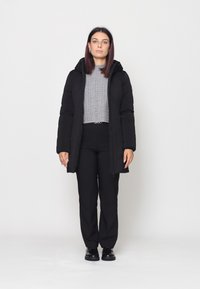 Giacca a vento nera con cappuccio, chiusura con zip, tasche laterali e un maglione in grisaglia grigia sotto, abbinata a pantaloni e scarpe neri.