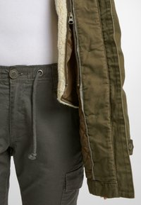 Armádně zelená užitková bunda s kožešinovou podšívkou, zipovým zapínáním a knoflíkovými detaily. Kombinovaná s tmavě zelenými kalhotami typu cargo se stahováním v pase na šňůrku.