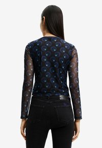 Blusa a maniche lunghe traslucida con motivi circolari blu su sfondo nero, abbinata a jeans neri a vita alta.