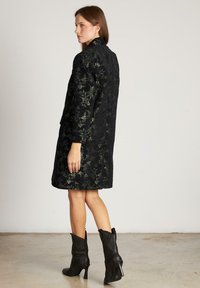 Mujer de pie con un abrigo de flores negro hasta la rodilla y botines negros de tacón contra un fondo claro y liso.