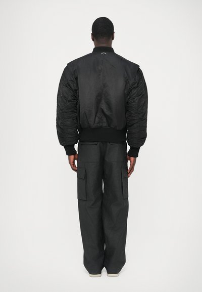 Juoda bombonešio striukė su dygsniuotais pečiais ir ribotais rankogaliais, derinama su laisvo kirpimo pilkomis kargo kelnėmis, turinčiomis kelias kišenes.