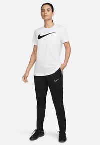 Weißes T-Shirt mit einem schwarzen Nike-Swoosh, kombiniert mit schwarzen Hosen, die ein kleines graues Nike-Logo aufweisen. Designed für sportliche Kleidung.