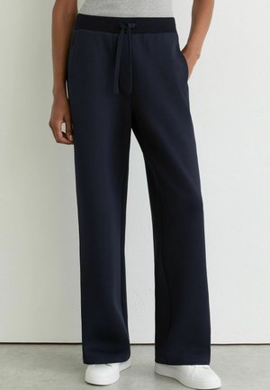 Persoon draagt zwarte wijde broek met elastische tailleband en trekkoord, gecombineerd met witte sneakers en een lichtgrijs topje.