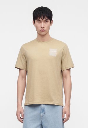 Junger Mann trägt ein beiges T-Shirt mit kurzen Ärmeln und kleinem weißen "Michael Kors"-Logo auf der Brust, kombiniert mit hellblauen Jeans.