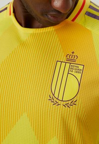 Gul sportsjersey med struktureret design, med et lilla emblem og striber. Inkluderer teksten "Royal Belgian FA 1895."