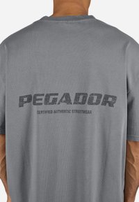 Tricou din bumbac gri, având un imprimeu negru mare, uzat, care spune "PEGADOR" și "CERTIFICAT AUTENTIC STREETWEAR" pe spate.