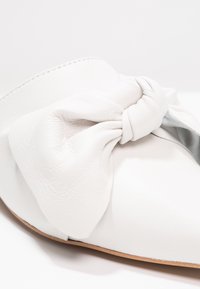 Gros plan d'une chaussure blanche en cuir à enfiler, présentant un grand nœud texturé sur la partie supérieure avec un bord de semelle marron clair.