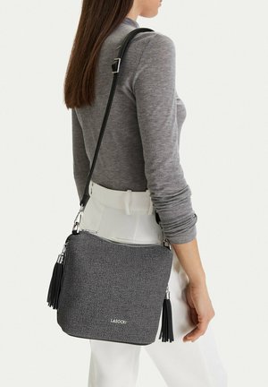 Mujer con top de manga larga gris y pantalones blancos, llevando un bolso bandolera gris texturizado con flecos y el logo de LASOCKI.