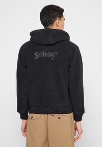 Schwarzer Kapuzenpullover aus Fleece-Material, mit geripptem Saum und Bündchen, einem strukturierten Logo auf dem Rücken, getragen über beige Hose.