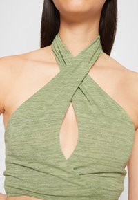 Top verde sem mangas feito de um tecido texturizado suave, com um design cruzado e um recorte em forma de gota no peito.