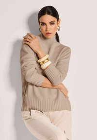 Frau mit dunklen Haaren, die zurückgebunden sind, trägt einen beigen Rollkragenpullover, helle Hosen, goldene Creolen und übereinander gestapelte Armbänder in neutralen Tönen.