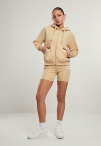 Felpa beige con zip e due tasche frontali, abbinata a pantaloni da ciclismo beige con dettaglio del logo. La modella indossa sneakers e calze bianche.