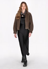 Bomber jacket in suede marrone con zip frontale, maniche oversize e un vestito di seta nera. La modella indossa stivaletti neri alla caviglia e una sciarpa a fantasia.