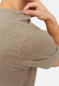 Beige t-shirt met korte mouwen, voorzien van een ronde halslijn en subtiele textuur, met een hand die de kraag aan de achterkant aanpast.