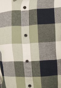 Groene en crèmekleurige geruite shirt met een groot ruitpatroon en donkere marineblauwe accenten; voorzien van zwarte knopen en een kraag.