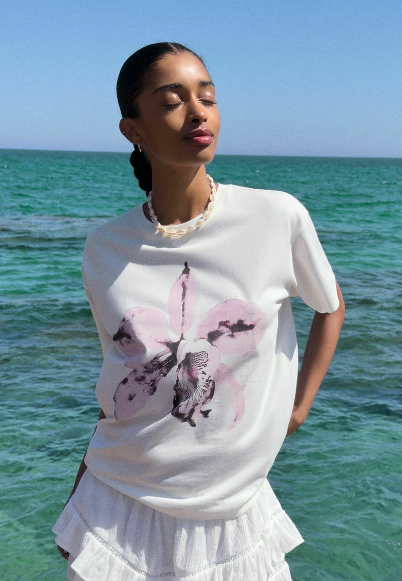 T-shirt en coton blanc avec un imprimé orchidée rose et noir, complété par un collier en coquillage texturé et une jupe blanche à volants superposés.