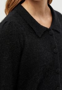 Svart stickad cardigan med krage, texturerad tyg och tre knappar. Materialet verkar mjukt och lätt fuzzy.