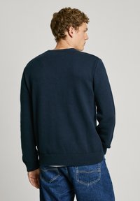 Navy blue knitted sweater with ribbed cuffs and hem, featuring a classic crew neckline and a textured finish. Worn with blue denim jeans.