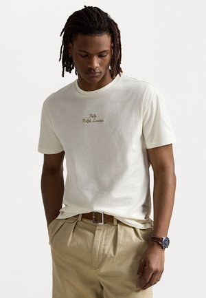 Kreemvärvi T-särk ümmarguse kaelusega, millel on pruuni niidiga brodüüritud "Polo Ralph Lauren". Sobitatud heledad beežid püksid ja pruun vöö.