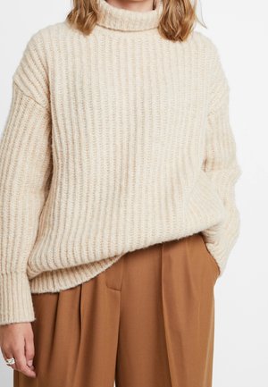 Maglione beige a collo alto a coste con una vestibilità rilassata, caratterizzato da un filato spesso e testurizzato e spalle scese, abbinato a pantaloni marroni plissettati.
