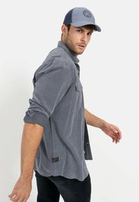 Graues Hemd mit langen Ärmeln und aufgerollten Ärmeln, mit einer Brusttasche und einem Etikett am Saum, kombiniert mit einer blauen Baseballmütze mit Logo.