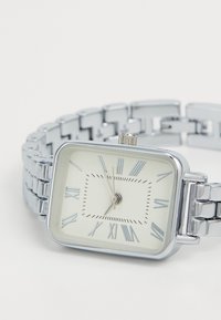 Silberne rechteckige Uhr mit einem weißen Zifferblatt, römischen Ziffern und einem metallischen Gliederband. Verfügt über eine Krone an der Seite zum Einstellen der Zeit.
