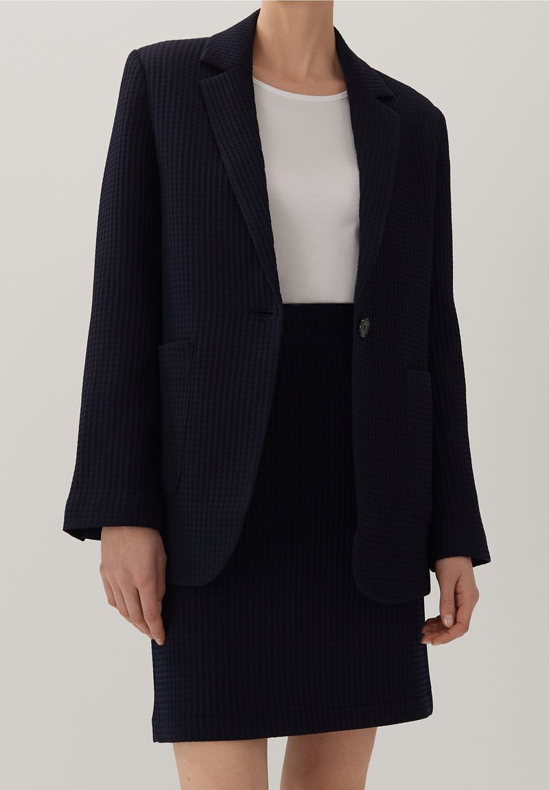 Femme portant un blazer noir texturé et une jupe assortie sur un haut blanc uni, debout devant un fond neutre clair.