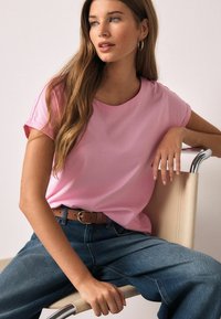 Tricou roz cu mâneci scurte și decolteu rotund, asortat cu blugi albastri din denim cu talie înaltă. O curea maro cu o cataramă aurie completează ținuta.