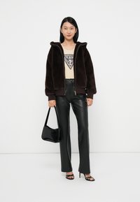 Veste bomber en fausse fourrure marron, tee-shirt graphique crème avec un design en diamant, pantalon en fausse cuir noir, sac à main noir et talons aiguilles noirs avec lanières.