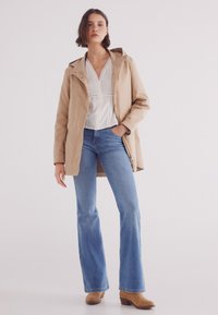 Cappotto impermeabile beige con cappuccio, chiusura a bottoni, indossato sopra una camicetta bianca e jeans a zampa blu. Stivaletti alla caviglia marroni. Tessuto morbido.