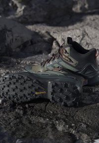 Botas de senderismo con suelas Continental resistentes descansando sobre terreno rocoso, con colores negro y verde y cordones con acentos naranjas.