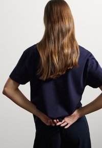 T-shirt in cotone blu navy con maniche corte, caratterizzato da un design rilassato. I capelli scendono in onde morbide lungo la schiena. Ambientazione minimalista.