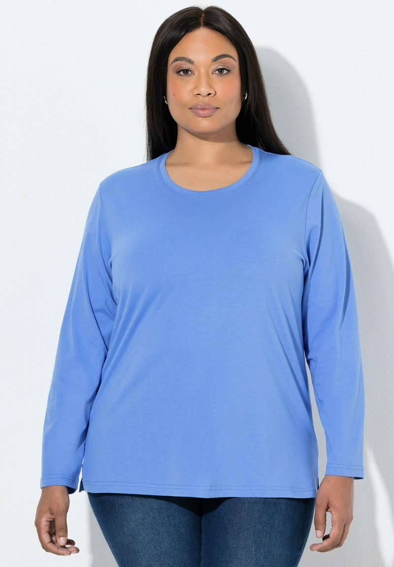 Vrouw met lang donker haar, gekleed in een lichtblauw shirt met lange mouwen en donkere spijkerbroek, staand tegen een egaal witte achtergrond.